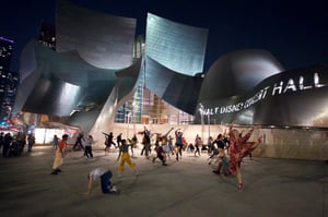 Nighttime scene in an outdoor courtyard at Walt Disney Concert Hall. A group of people with varied appearances—different ages, skin tones, and styles of dress—are gathered together wearing casual clothing. They are dancing in a modern, expressive way, with arms and bodies moving freely rather than in a set pattern. Behind them, the curved metal exterior of the concert hall is lit from below, casting soft, bright light upward and creating a glowing backdrop. The dancers are spread across the open space, and the lighting highlights their movement against the darker night sky.
