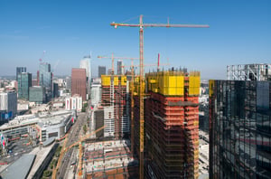 View of the South Park neighborhood in downtown Los Angeles. Several tall buildings fill the scene, including multiple skyscrapers under construction. Construction cranes are positioned above the buildings, with long horizontal arms extending across the sky. Some structures appear unfinished, with exposed frameworks or partially completed exteriors. The scene shows an active development area, with the cranes and incomplete buildings indicating ongoing construction.