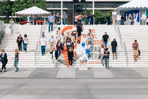 Daytime scene at the steps of The Music Center. A group of people with varied appearances—different ages, skin tones, and clothing styles—are walking down a wide set of steps. The steps have the words “The Music Center” written across them in large letters. The people are spread out across the steps, some walking alone and others in small groups. The scene shows an active public space with people moving downward in natural, everyday motion.