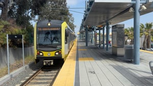An above-ground light rail station in Los Angeles with a stopped train on the tracks. The front of the train displays its destination, “Santa Monica,” on a digital sign. The train is part of the E Line. The platform runs alongside the train, with space for passengers to wait and board. Overhead structures and rail lines are visible, indicating an outdoor transit stop.