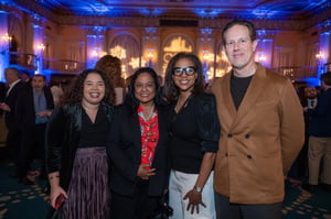 A group of four adults stands together at a formal indoor event in a large, ornate ballroom lit with blue and warm ambient lighting. From left to right: a woman with curly hair wearing a black top and a pleated skirt smiles at the camera; next to her is Los Angeles City Councilmember Heather Hutt, wearing a black blazer over a red printed blouse, also smiling; beside her, another woman in a black top with glasses and light-colored pants smiles; and on the far right, a man in a tan blazer stands with his hands behind his back, smiling. Other guests are visible in the softly blurred background, mingling beneath decorative architectural details and chandeliers.