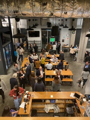 A large group of adults gathers at a CCA member mixer in an open, industrial-style venue with exposed ductwork and high ceilings. Attendees stand and sit around long wooden tables and benches, talking in small groups, holding drinks, and networking. In the foreground, a bar counter with bottles and glassware is visible, while in the middle of the room people mingle and converse. Toward the back, additional guests stand near high-top tables and a wall with an illuminated exit sign. The atmosphere is social and professional, reflecting a networking event hosted by the CCA, an organization that brings together business and community leaders in Los Angeles to foster connections and collaboration.