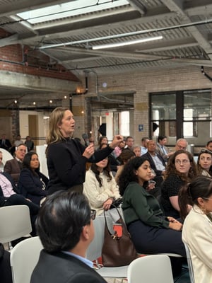A woman stands among seated attendees at an indoor event in an industrial-style space with exposed brick walls and visible ceiling beams. She holds a microphone in one hand and gestures with the other as she speaks. She wears a dark long-sleeve top and has shoulder-length light brown hair. Around her, a diverse group of adults sits in rows of chairs, facing her and listening; some look directly at her while others glance toward the front of the room. As a member of CCA, she is actively participating in a Q&A session with a policymaker.