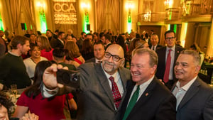 A group of attendees gathers for a photo at the Central City Association’s holiday party in a large, warmly lit ballroom. Los Angeles Police Department Chief Jim McDonnell, wearing a green tie, stands at center smiling as a man beside him holds up a phone to take a selfie. Other guests in business attire fill the background, mingling and conversing, with CCA branding visible on the wall behind them.