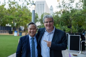 Los Angeles City Councilmembers John Lee (left) and Tim McOsker (right) stand together outdoors at the Central City Association’s “Elected Officials” event. They smile toward the camera, with McOsker placing an arm around Lee and gesturing toward him. Trees, lawn, and civic buildings are visible in the background, reflecting a relaxed, outdoor event setting.