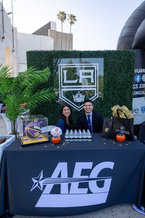 Two people sit behind a branded table at a sponsored activation during the Central City Association’s “Treasures of Los Angeles” event. They smile toward the camera while seated in front of a green backdrop featuring the LA Kings logo. The table displays promotional items, including bottled water, a basketball in a clear case, and small giveaway items. Additional branded signage and materials are arranged around the booth, indicating a sponsor presence. The activation is set outdoors, with buildings and palm trees visible in the background.