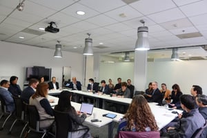 A group of about twenty adults sits around a U-shaped arrangement of white tables during a CCA policy committee meeting in a modern conference room. Attendees wear business or business-casual attire and face inward toward the discussion. Several people take notes on laptops or paper, while others listen attentively. On the right side of the room, a man in a suit appears to be speaking as others look in his direction. Overhead lights illuminate the space, and a wall-mounted screen and glass partition are visible in the background. The setting conveys a formal meeting focused on discussion and collaboration.
