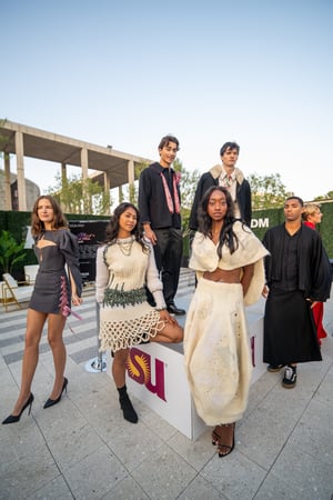 Models of diverse races and genders pose in Arizona State University student-designed outfits, showcasing different styles and fabrics at the Music Center as part of the Central City Association’s “Treasures of Los Angeles” event. They wear a variety of The setting is a staged event space with theatrical lighting, emphasizing the creativity and celebratory atmosphere of the program.