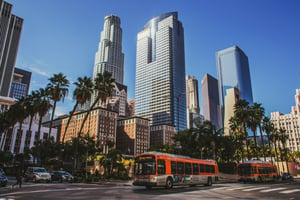 Daytime view of a downtown Los Angeles street. An orange city bus from Los Angeles Metro is traveling along the road. Tall buildings line both sides of the street, including multiple skyscrapers. In the background, the rear side of the Millennium Biltmore Hotel is visible near Pershing Square. The scene is viewed at street level, showing the bus moving through an active urban area.