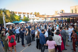 Outdoor daytime gathering in the courtyard of LA Plaza de Cultura y Artes. Hundreds of people dressed in summer business attire are standing, walking, and mingling in groups. The courtyard is open-air, with historic architecture surrounding the space. The atmosphere is lively and social, with attendees engaged in conversation throughout the area. The scene conveys a professional networking or cultural event in a public courtyard setting.